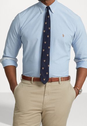Mann trägt ein hellblaues Hemd mit Knöpfen und aufgerollten Ärmeln, eine marineblaue gemusterte Krawatte, beige Hose, braunen Ledergürtel und Armbanduhr.