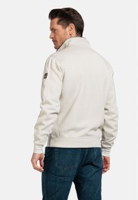Lichtcrème sweatshirt met een hoge kraag, ribgebreide boorden en onderrand, met een zwart logo label op de linkermouw. Draagt met blauwe jeans.