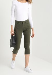 Pantalones cortos en algodón elástico color verde oliva, combinados con una blusa blanca de manga larga y zapatillas blancas, sosteniendo un bolso negro en una mano.