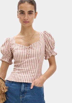 Kvinde iført en lyserød og hvid lodret striped puff sleeve top med en lille sløjfe foran, blå jeans, et gyldent halskæde og holder en frynset taske.