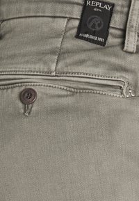 Jeans de mezclilla gris claro con un bolsillo cosido, cremallera de metal y una etiqueta negra de marca. La textura es suave con un ligero brillo.
