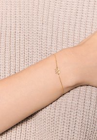 Złota bransoletka o minimalistycznym wzornictwie, z centralnym charms'em w kształcie litery M, opierająca się na nadgarstku na teksturowanym, beżowym tle z dzianiny.