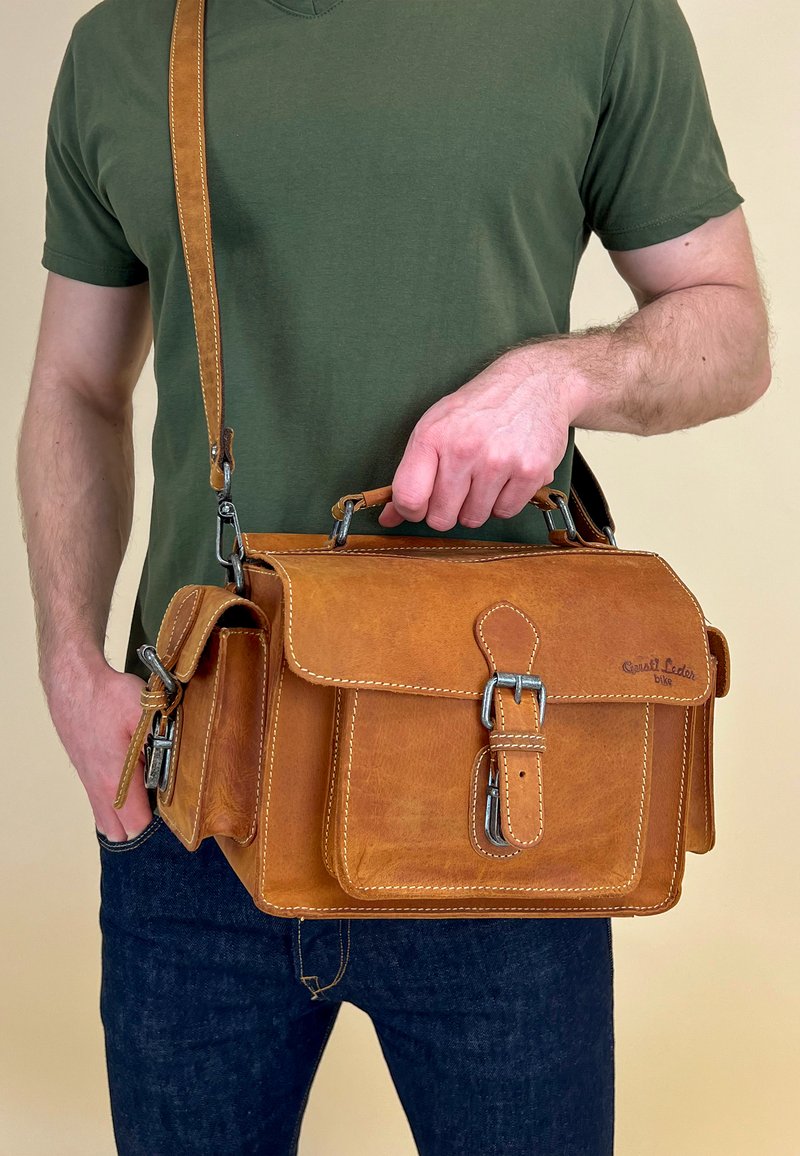 Brun læder messenger-taske med lukkeflap, metalspænde og skulderrem. Indeholder en sidelomme og synlige syninger.