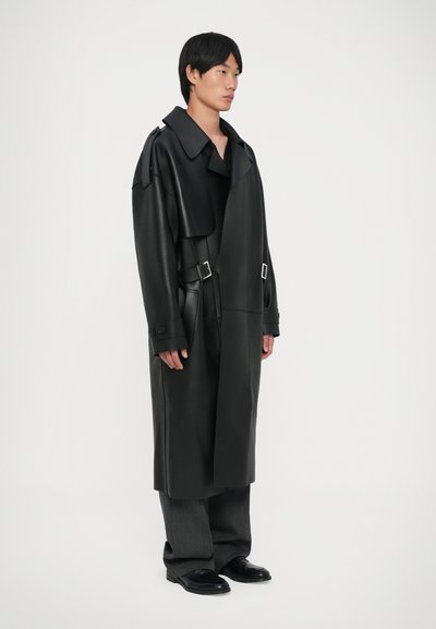 Sort læder trench coat med en stor krave, justerbare side spænder og knapmanchetter, parret med grå brede bukser og sorte sko.