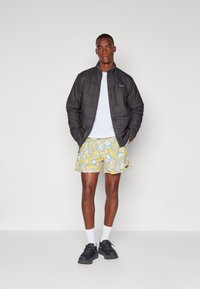 Veste matelassée noire, t-shirt blanc, shorts à motifs jaunes, baskets noires, chaussettes blanches. Modèle debout devant un fond neutre.