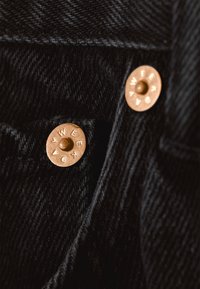 Zwarte denimstof met een gestructureerd weefsel. Voorzien van twee koperen knopen met gegraveerde letters, dicht bij elkaar geplaatst op de tailleband.