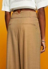 Beige tailored trousers with a high waist, back pockets, textured fabric, and a V-shaped cut at the waistband.