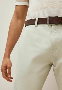 Chinos beige avec une texture lisse, accompagnés d'une ceinture en cuir marron avec une boucle en argent. Couture visible et design ajusté.