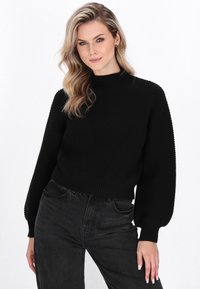 Sort ribbet sweater med høj krave, der har ballonsleeves og et cropped design, kombineret med højtaljede mørke jeans.