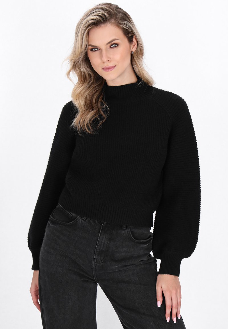 Sort ribbet sweater med høj krave, der har ballonsleeves og et cropped design, kombineret med højtaljede mørke jeans.
