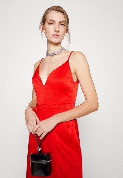Abito di raso rosso con scollatura a V profonda e spalline sottili, abbinato a una piccola borsa in vernice nera con zip e un manico arrotondato.