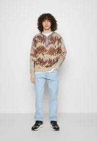 Suéter de punto multicolor con escote en V y media cremallera, que presenta patrones abstractos en tonos marrones, verdes y morados, combinado con jeans de color azul claro.
