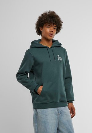 Jeune homme aux cheveux bouclés portant un sweat à capuche vert foncé avec un motif graphique de geste de main blanche et un jean bleu clair, debout avec une main dans la poche.