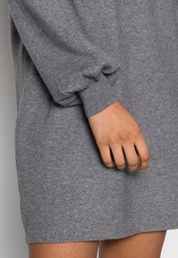 Main de femme à côté de l'ourlet et du poignet d'une robe-pull grise en maille, montrant le détail côtelé du poignet et la texture douce du tissu.