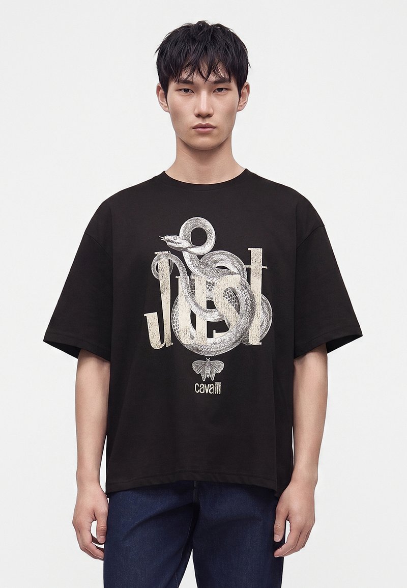 Νέος άνδρας με μαύρο T-shirt που έχει μεγάλο λευκό γραφικό φιδιού και τη λέξη "Just" στο μπροστινό μέρος, συνδυασμένο με σκουρόχρωμο παντελόνι.