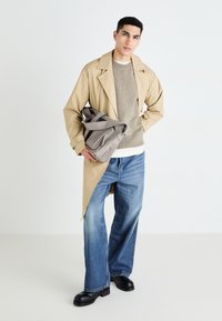 Un manteau trench beige, un pull gris, un jean bleu large et des chaussures noires. Le modèle tient un sac gris avec des doubles zippers et des anses. Textures lisses.