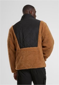 Smeđi krzneni jakna s crnim kvadratnim panelom na leđima, s visokim ovratnikom i elastičnim manžetama za dodatnu toplinu i udobnost.