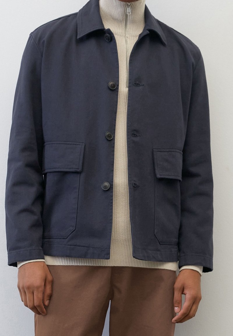 Homme portant une veste bleu marine boutonnée par-dessus un pull beige zippé avec un pantalon marron, debout devant un fond blanc uni.