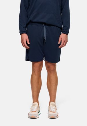 Hombre vistiendo pantalones cortos azul marino, sudadera con capucha azul marino a juego y zapatillas beige, de pie frente a un fondo blanco liso.