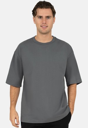 Junger Mann mit kurzen braunen Haaren, der ein schlichtes, übergroßes graues T-Shirt trägt und mit einer Hand in der Tasche vor einem weißen Hintergrund steht.