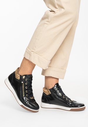 Legs wearing beige corduroy pants and shiny black high-top sneakers with gold accents and white soles, standing on a white background.