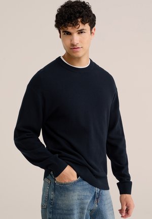 Joven con cabello rizado y oscuro, vestido con un suéter azul marino con textura y vaqueros azul claro, posando con una mano en el bolsillo sobre un fondo neutro.