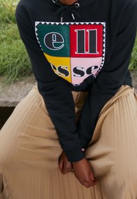 Černá mikina s barevnou grafikou obsahující zelené, červené, žluté a růžové čtverce. Plisované béžové kalhoty z texturované látky, sedící pozice.