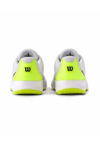 Zapatillas deportivas blancas con acentos en el talón de un amarillo brillante y suelas de goma texturizadas. Presentan un sutil logo de la marca en la parte trasera.