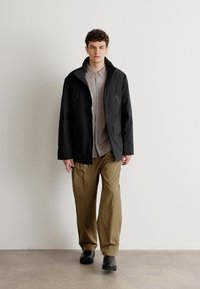 Giacca impermeabile nera con cappuccio, due tasche con zip, indossata sopra una camicia a bottoni grigia chiara e pantaloni khaki. Finitura opaca e vestibilità rilassata.