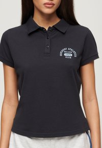 Polo de color azul marino oscuro con cuello, que presenta un logo en el pecho 'Superdry Athletics Beach 1979 Club' en bordado azul claro y un diseño ajustado.