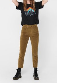 Pantalon en velours côtelé marron à coupe slim, avec une taille haute, cinq poches, et une longueur cheville, assorti à un t-shirt graphique noir et des bottes noires.