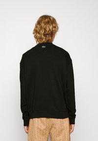 Sudadera negra de corte relajado y puños acanalados, con un pequeño logo verde de Lacoste en la parte posterior. Diseño simple y de color sólido.