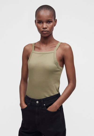 Jeune personne aux cheveux courts portant un débardeur vert olive ajusté et un pantalon noir, debout les mains dans les poches devant un fond uni.