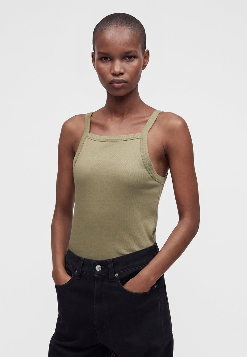 Jeune personne aux cheveux courts portant un débardeur vert olive ajusté et un pantalon noir, debout les mains dans les poches devant un fond uni.