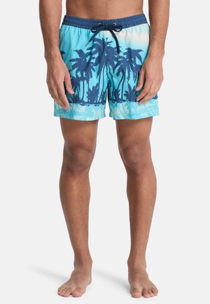Mann trägt blaue Badeshorts mit dunkelblauem Palmenmuster, steht barfuß vor einem weißen Hintergrund.