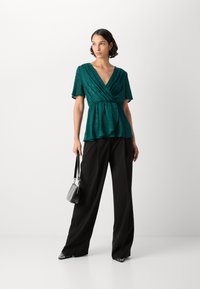 Top avvolgente teal con maniche corte e orlo a peplo, abbinato a pantaloni neri a gamba larga e una piccola borsa nera. Tessuto di texture liscia.