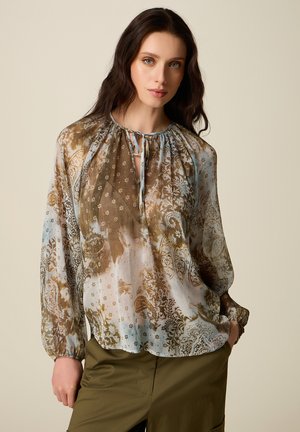 Donna con lunghi capelli scuri che indossa una camicetta trasparente con motivo floreale nei toni del marrone e del blu e pantaloni verde oliva, con le mani parzialmente nelle tasche.