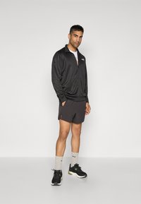 Sort zip-up jakke med krave, grå skjorte, mørke shorts, grå sokker og sorte sneakers. Glat stof, afslappet pasform, minimalistisk design.