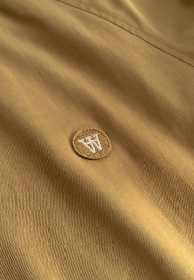 Tissu marron clair avec une petite écusson rond présentant un logo "W" blanc brodé près d'une couture.
