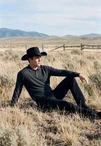 Homme portant un chapeau de cow-boy noir et des vêtements foncés assis sur une prairie sèche avec des montagnes et une clôture en bois en arrière-plan.