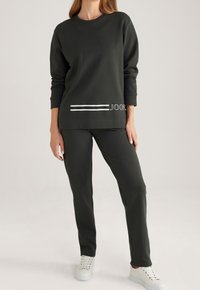 Tmavě zelená mikina a kalhoty ve volném střihu. Mikina má bílé vodorovné pruhy a logo na lemu. Bílé tenisky.