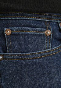 Dark blue denim jeans pocket with orange stitching and two copper rivets engraved with "JACK & JONES" on a black shirt background.