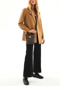 Borsa a tracolla marrone con design a motivo, hardware in oro e una lunga tracolla, abbinata a un blazer beige e pantaloni neri.