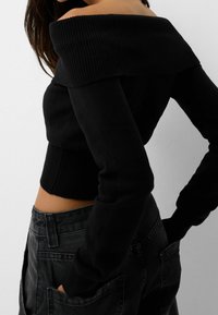 Suéter negro de hombros descubiertos con cuello acanalado y mangas largas, combinado con jeans oscuros que tienen grandes bolsillos y un aspecto desgastado.