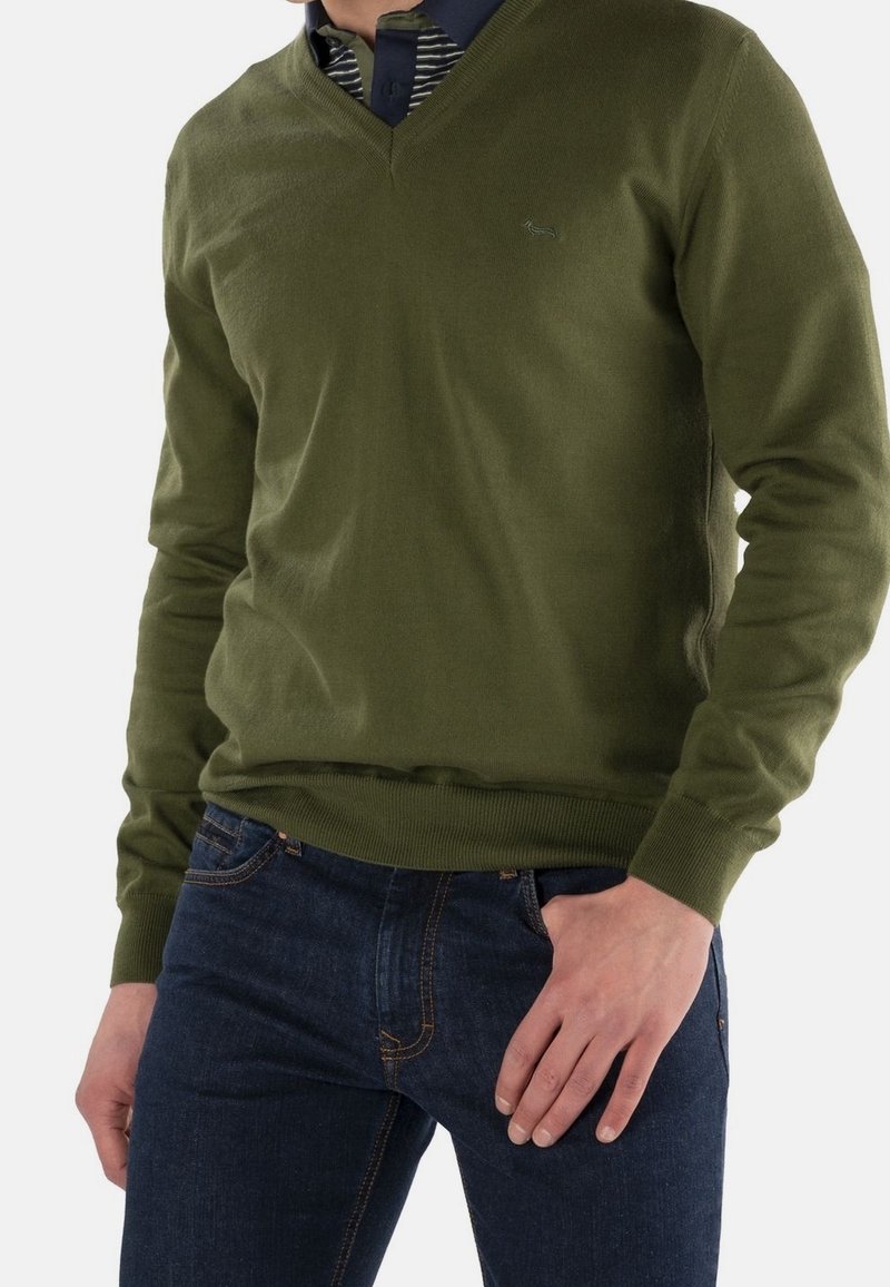 Oliwkowy sweter v-neck wykonany z miękkiej dzianiny. Posiada prążkowane mankiety i dół. Noszony na niebiesko-paskowanej koszuli, w połączeniu z ciemnymi dżinsami.