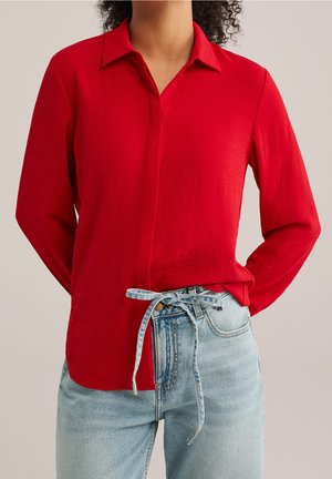 Vrouw draagt een rode blouse met lange mouwen en knopen, ingestopt in lichtblauwe spijkerbroek met een geknoopt denimriempje in de taille.