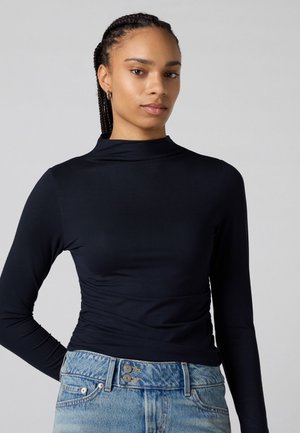Haut à col roulé bleu marine à manches longues, fabriqué en tissu lisse et extensible, avec une silhouette ajustée et un léger ruché à la taille.