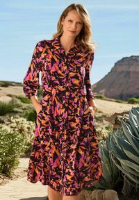 Blumengemustertes Kleid mit langen Ärmeln, Knöpfen und tailliertem Schnitt. Die Farben umfassen Pink, Orange und Schwarz. Verfügt über Taschen und einen ausgestellten Rock.