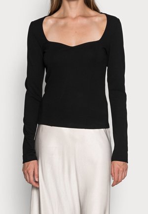 Mujer con una blusa negra de manga larga y escote cuadrado, y una falda blanca de satén de talle alto, frente a un fondo liso.