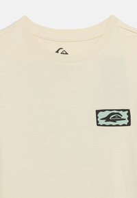 Μπεζ μπλούζα με στρογγυλή λαιμόκοψη και ένα μικρό ορθογώνιο λογότυπο σε τιρκουάζ και μαύρο στο πάνω αριστερό μέρος του στήθους.